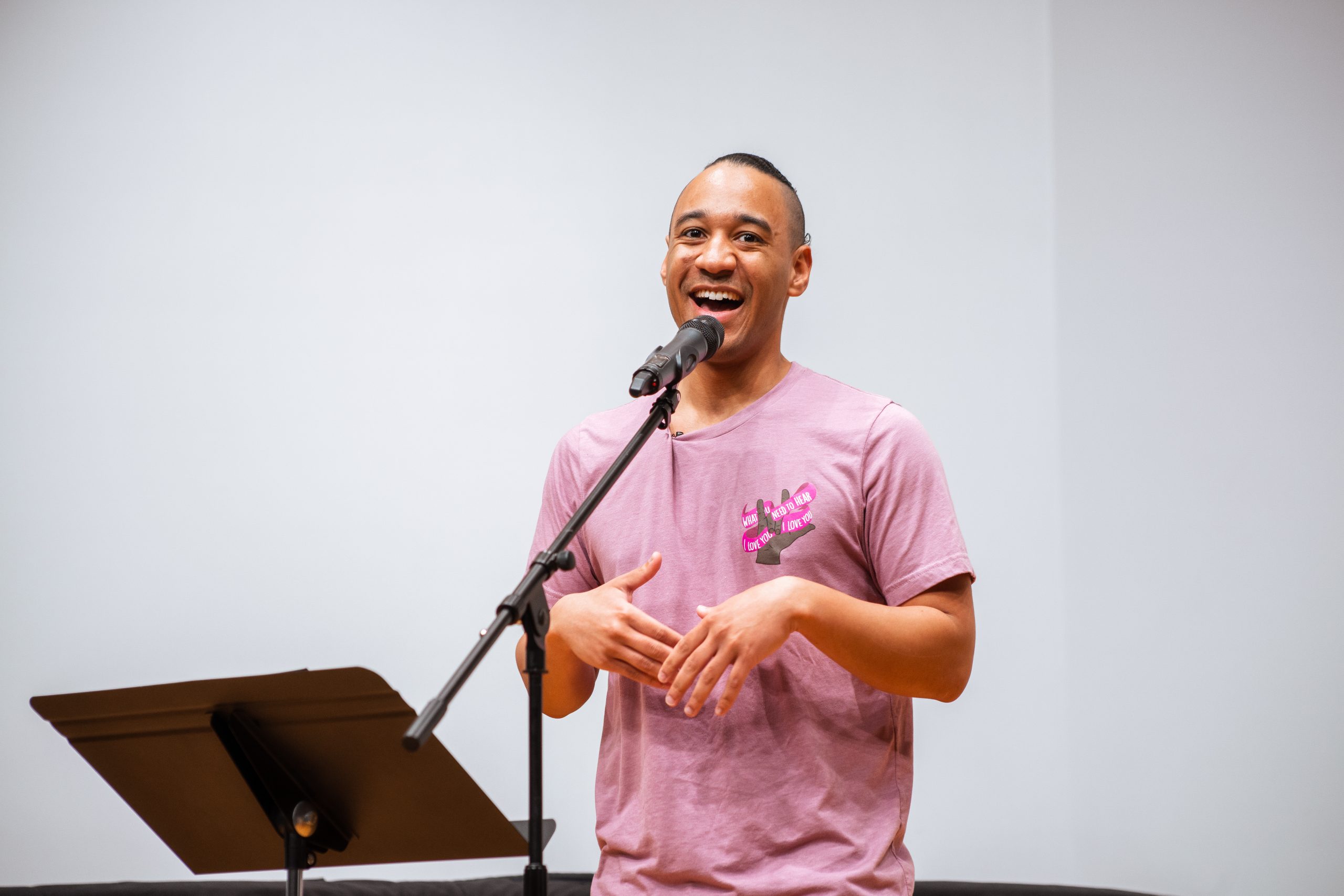 Portrait of a person in a pink t-shirt smiling while speaking into a microphone in front of a podium.