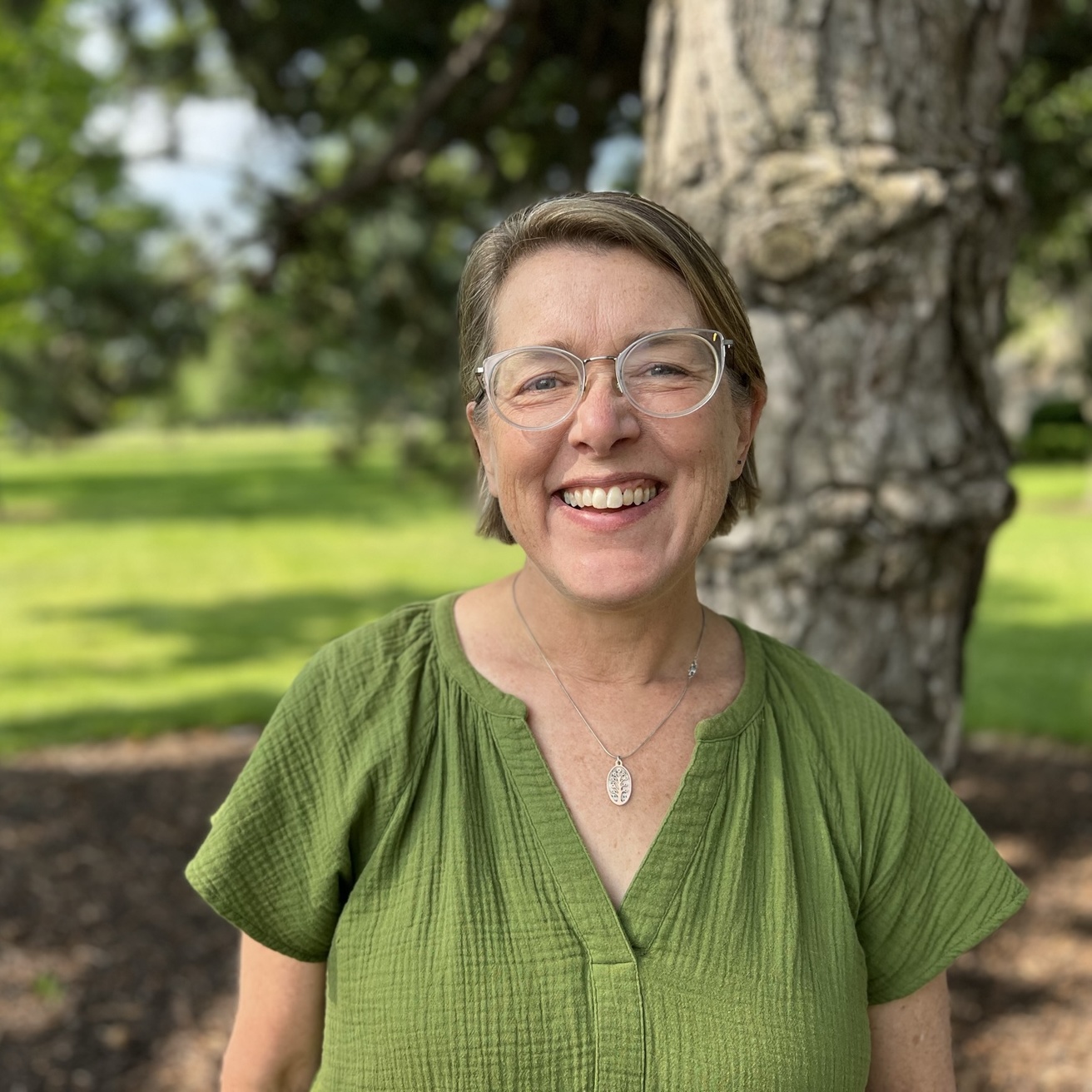 Portrait of a woman in a green shirt with clear-framed glasses smiling in front of a tree.