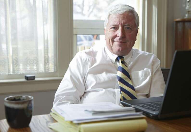 Portrait of a man with a striped tie and a white button-up in front of a window. There is a desk in front of him with a laptop and stack of papers.