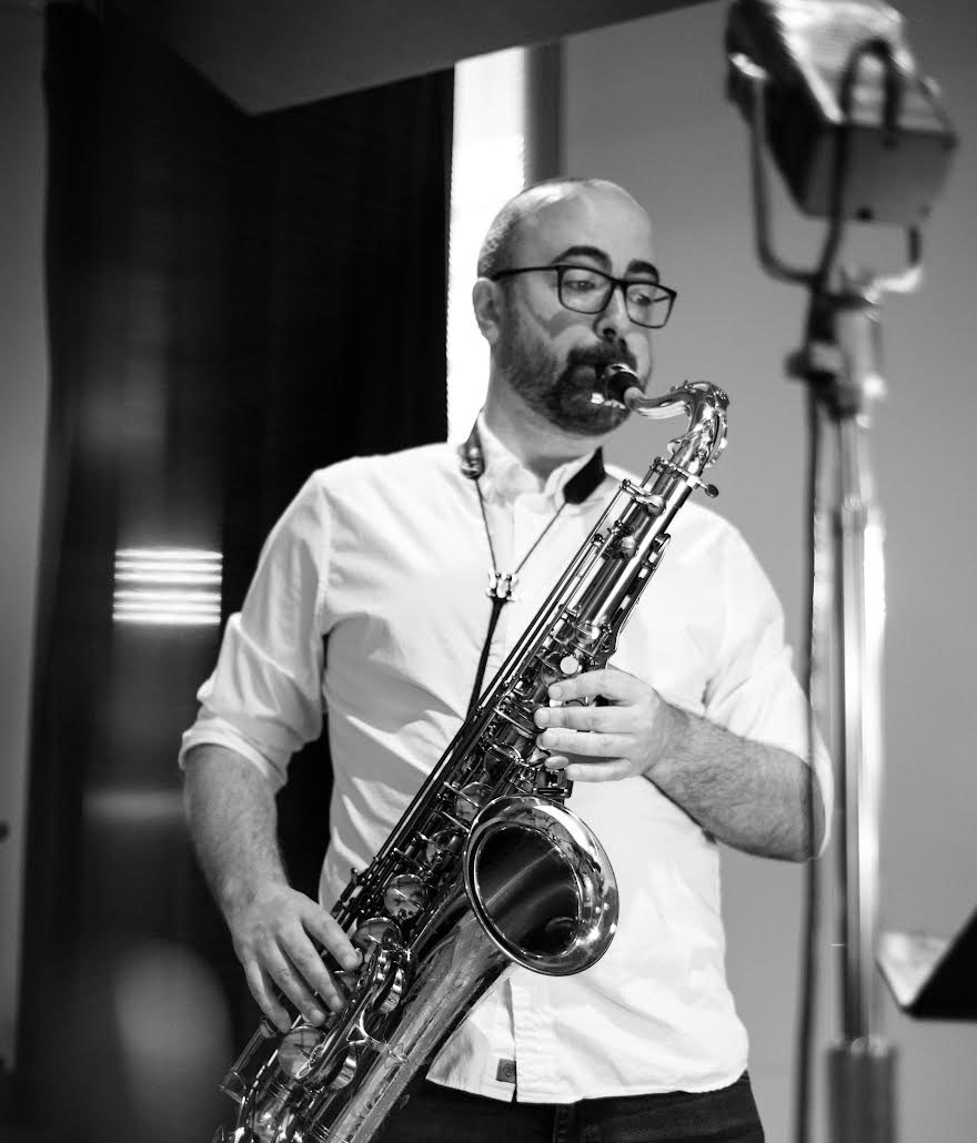 Black and white portrait of a person with glasses in a button-up shirt playing saxophone.