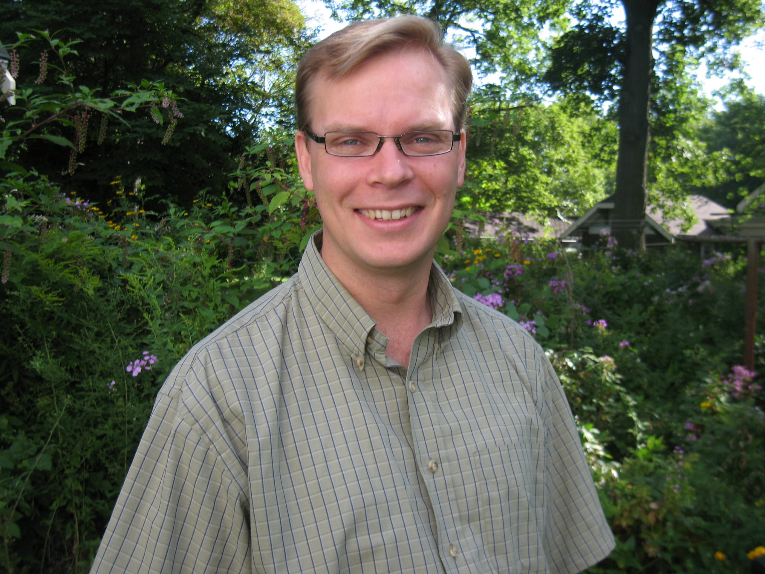 Head and shoulders portrait of a man with glasses, short brown hair, and a tan, checkered shirt smiling in front of foliage.