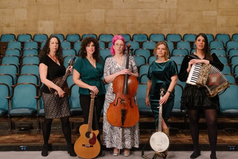 A group of five women Americana musicians standing together and holding their instruments in front of rows odf empty theatre seats